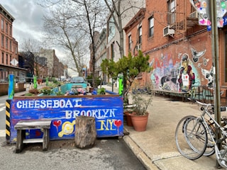 A colorful street scene in Brooklyn featuring a vibrant mural on a brick wall depicting a mix of urban and natural imagery, including birds and graphic elements. In the foreground, a decorative barrier made of blue-painted wood with 'CHEESEBOAT BROOKLYN' in white letters serves as a marker for a small outdoor seating area. Potted plants, a tree, and rustic seating are placed around it. A bicycle is parked nearby, and buildings line the street, which stretches into the background.