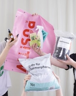 Several hands are holding various gardening and plant care products against a light background. These include a pink banner with bold red text, a bottle of plant spray, gardening tags, bottles of liquid fertilizer, and bags of plant soil labeled in German.