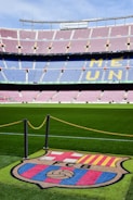 A football stadium with tiered seating in red and blue colors, featuring a yellow text spelling out 'ME UNIC' on the seats. The foreground shows a vibrant green football pitch and a large emblem with 'FCB' on it, surrounded by a chain.