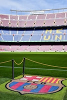 A football stadium with tiered seating in red and blue colors, featuring a yellow text spelling out 'ME UNIC' on the seats. The foreground shows a vibrant green football pitch and a large emblem with 'FCB' on it, surrounded by a chain.