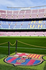A football stadium with tiered seating in red and blue colors, featuring a yellow text spelling out 'ME UNIC' on the seats. The foreground shows a vibrant green football pitch and a large emblem with 'FCB' on it, surrounded by a chain.