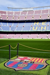 A football stadium with tiered seating in red and blue colors, featuring a yellow text spelling out 'ME UNIC' on the seats. The foreground shows a vibrant green football pitch and a large emblem with 'FCB' on it, surrounded by a chain.