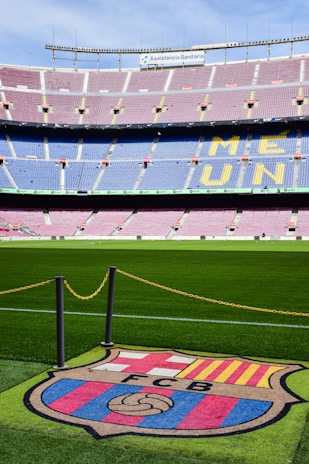 A football stadium with tiered seating in red and blue colors, featuring a yellow text spelling out 'ME UNIC' on the seats. The foreground shows a vibrant green football pitch and a large emblem with 'FCB' on it, surrounded by a chain.