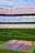 A football stadium with tiered seating in red and blue colors, featuring a yellow text spelling out 'ME UNIC' on the seats. The foreground shows a vibrant green football pitch and a large emblem with 'FCB' on it, surrounded by a chain.