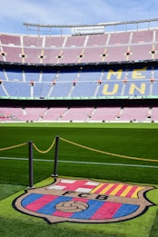A football stadium with tiered seating in red and blue colors, featuring a yellow text spelling out 'ME UNIC' on the seats. The foreground shows a vibrant green football pitch and a large emblem with 'FCB' on it, surrounded by a chain.