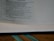 A close-up view of an open book, showing text on a page. The book is lying flat with a visible binding, and lines of text are seen clearly on the page. There is a ribbon bookmark with two green strands visible at the bottom of the image.