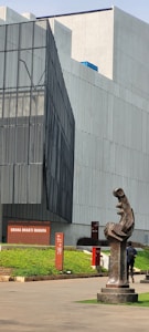 A modern architectural building with angular lines and sleek, large windows. In the foreground, there is a dark abstract sculpture that stands on a pedestal. The building has a sign reading 'GRAHA BHAKTI BUDAYA' and is surrounded by neatly trimmed greenery. A person is walking near the sculpture.