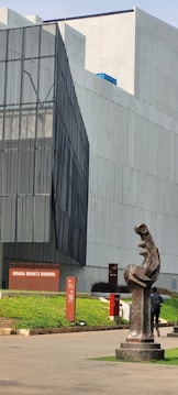 A modern architectural building with angular lines and sleek, large windows. In the foreground, there is a dark abstract sculpture that stands on a pedestal. The building has a sign reading 'GRAHA BHAKTI BUDAYA' and is surrounded by neatly trimmed greenery. A person is walking near the sculpture.