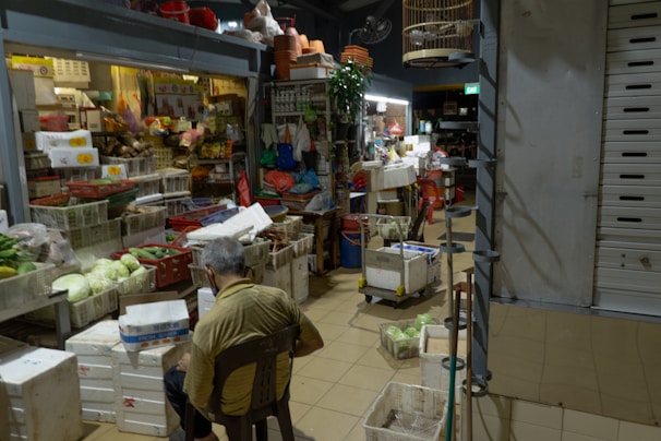 An indoor market stall filled with various goods, including fresh vegetables, packaged food items, and household products. The stall is crowded with boxes, crates, and baskets. A man, seated with his back to the camera, is wearing a mask and seems to be taking a break or organizing items. The space has subdued lighting with a ceiling fan and hanging birdcage visible.