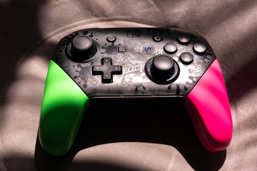 A video game controller featuring a black central section with intricate designs and buttons, flanked by vibrant green and pink grips. It is illuminated dramatically with light and shadow creating a dynamic texture across the surface.