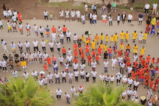 A large group of people is organized in rows, wearing brightly colored uniforms, likely engaging in a coordinated activity such as exercise or training. The majority are in white, with distinct groups in red, yellow, and other colors. The setting appears to be outdoors on a paved surface with some greenery visible.