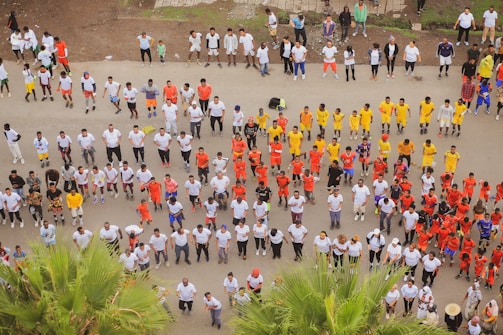 A large group of people is organized in rows, wearing brightly colored uniforms, likely engaging in a coordinated activity such as exercise or training. The majority are in white, with distinct groups in red, yellow, and other colors. The setting appears to be outdoors on a paved surface with some greenery visible.