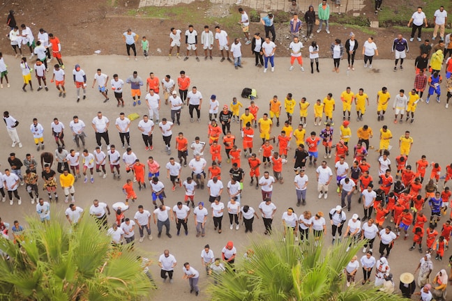 A large group of people is organized in rows, wearing brightly colored uniforms, likely engaging in a coordinated activity such as exercise or training. The majority are in white, with distinct groups in red, yellow, and other colors. The setting appears to be outdoors on a paved surface with some greenery visible.