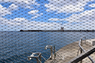 A scenic view of the ocean with a distant pier and a crane, seen through a wire mesh fence. The sky is partly cloudy with blue skies peering through. In the foreground, several security cameras are mounted on a concrete pathway beside the fence.