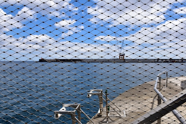 A scenic view of the ocean with a distant pier and a crane, seen through a wire mesh fence. The sky is partly cloudy with blue skies peering through. In the foreground, several security cameras are mounted on a concrete pathway beside the fence.