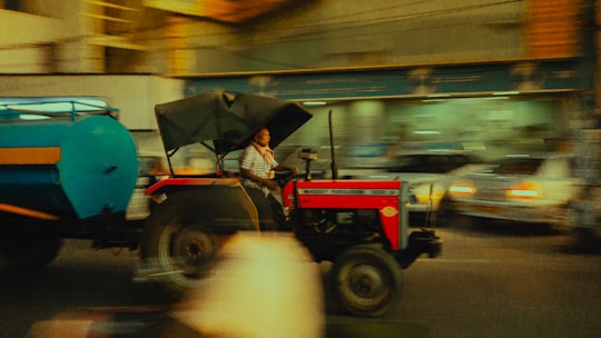 A person is driving a tractor with a large blue tank attached to it, moving through an urban area. The background is motion-blurred, indicating movement and speed. The tractor is red and has a canopy, while the surroundings include blurred images of cars and buildings.