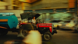 A person is driving a tractor with a large blue tank attached to it, moving through an urban area. The background is motion-blurred, indicating movement and speed. The tractor is red and has a canopy, while the surroundings include blurred images of cars and buildings.