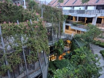 An urban environment featuring a multi-story building partially covered with lush green plants and ivy. The building has a modern architectural style with a blending of nature and structure. Walkways and balconies are visible, alongside trees and other vegetation in surrounding areas. The upper section of the building has a red-tiled roof with skylights.
