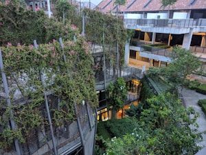 An urban environment featuring a multi-story building partially covered with lush green plants and ivy. The building has a modern architectural style with a blending of nature and structure. Walkways and balconies are visible, alongside trees and other vegetation in surrounding areas. The upper section of the building has a red-tiled roof with skylights.