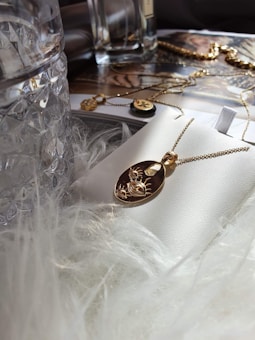 A gold pendant necklace with an engraved design rests on a white surface, surrounded by luxurious elements like a glass bottle and soft, fluffy material. In the background, there are more jewelry pieces and a magazine page with glamorous imagery.