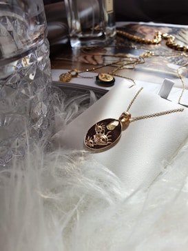 A gold pendant necklace with an engraved design rests on a white surface, surrounded by luxurious elements like a glass bottle and soft, fluffy material. In the background, there are more jewelry pieces and a magazine page with glamorous imagery.