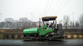 A green asphalt paver machine is parked on the side of a road. It has a large hopper at the front for asphalt material and a canopy covering the operator's seat. In the background, there are bare trees and a highway overpass. Buildings are visible in the distance under an overcast sky.