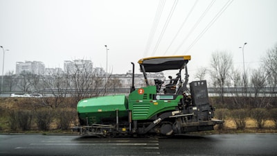 A green asphalt paver machine is parked on the side of a road. It has a large hopper at the front for asphalt material and a canopy covering the operator's seat. In the background, there are bare trees and a highway overpass. Buildings are visible in the distance under an overcast sky.