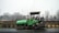 A green asphalt paver machine is parked on the side of a road. It has a large hopper at the front for asphalt material and a canopy covering the operator's seat. In the background, there are bare trees and a highway overpass. Buildings are visible in the distance under an overcast sky.