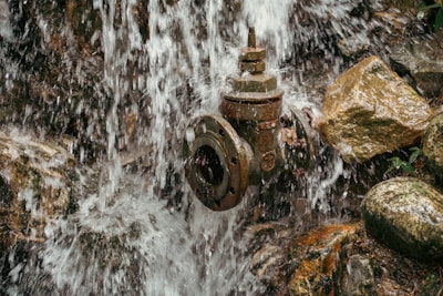 Water cascades down rocks, surrounding an old, rusted fire hydrant. The scene includes wet stones varying in size and shape, covered with trickling streams of water, creating a natural and textured environment.