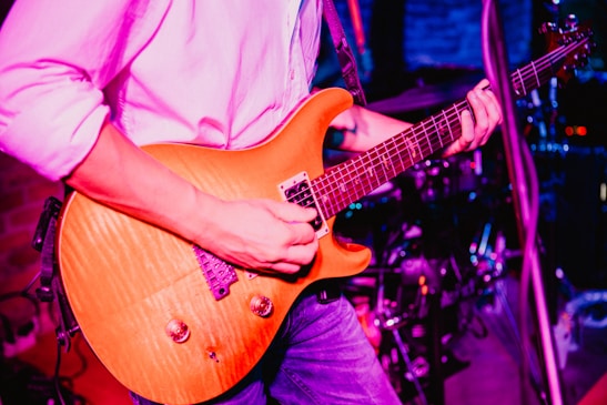 A person is playing an electric guitar with a light wood finish. The musician is wearing a white shirt and blue jeans. The background includes drum equipment and stage lighting, suggesting a live music performance.