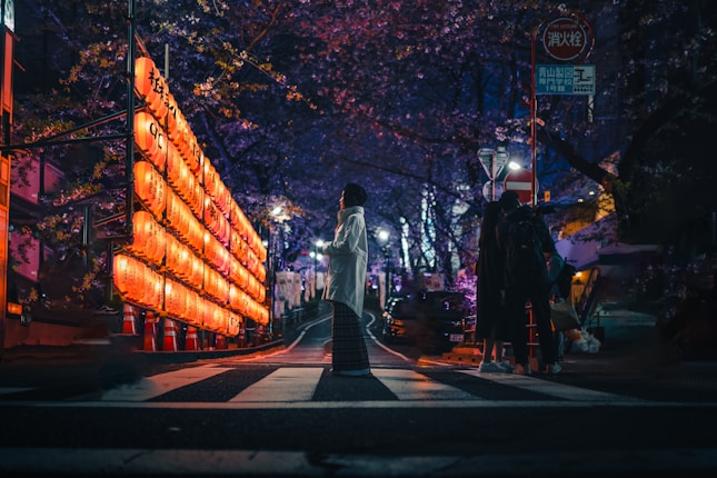 A street scene at night features a glowing row of orange lanterns lining the left side, casting a warm light on the surroundings. The road is lined with cherry blossom trees, creating an atmospheric canopy above. A person stands in the middle, facing the row of lanterns, while a group of people is visible to the right, casting long shadows.