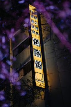 A brightly illuminated yellow sign with Japanese characters, mounted on the side of a building. The building appears to be modern, with visible windows. In the foreground, there are out-of-focus branches with purple flowers, adding a natural contrast to the urban setting.
