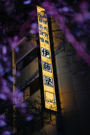 A brightly illuminated yellow sign with Japanese characters, mounted on the side of a building. The building appears to be modern, with visible windows. In the foreground, there are out-of-focus branches with purple flowers, adding a natural contrast to the urban setting.