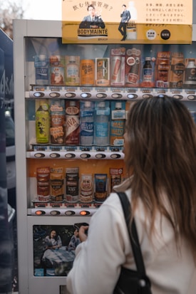 A person stands in front of a beverage vending machine stocked with various drinks, including canned coffee and bottled water. Several brands and colorful labels are visible, and there's an advertisement at the top with text and a photo of a person. The person appears to be making a selection, holding a wallet or phone.