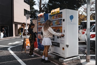 Two people are interacting with vending machines on a city street. The area is surrounded by cars and buildings, with trees visible in the background. One individual is using a white vending machine labeled 'Otsuka,' while the other is standing nearby looking at a colorful machine.