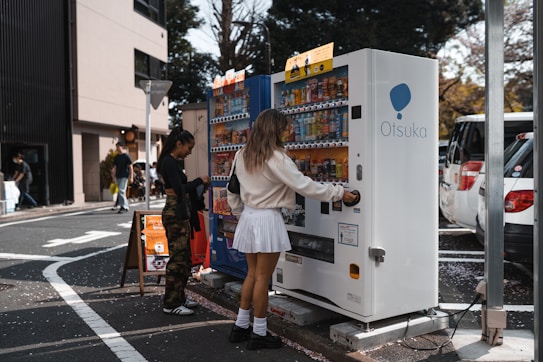 Two people are interacting with vending machines on a city street. The area is surrounded by cars and buildings, with trees visible in the background. One individual is using a white vending machine labeled 'Otsuka,' while the other is standing nearby looking at a colorful machine.