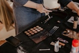 A person is applying makeup using a variety of tools on a black-draped table. There are makeup brushes, an eyeshadow palette, foundation bottle, and other cosmetic items scattered around. The scene suggests a makeup session or preparation for an event.