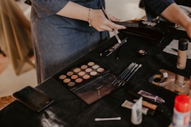 A person is applying makeup using a variety of tools on a black-draped table. There are makeup brushes, an eyeshadow palette, foundation bottle, and other cosmetic items scattered around. The scene suggests a makeup session or preparation for an event.