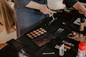 A person is applying makeup using a variety of tools on a black-draped table. There are makeup brushes, an eyeshadow palette, foundation bottle, and other cosmetic items scattered around. The scene suggests a makeup session or preparation for an event.