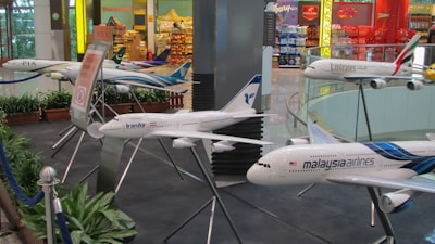 The image features a display of multiple model airplanes situated indoors. Various airline logos are visible on the models, including Malaysia Airlines, Emirates, IranAir, and PIA. The setting appears to be a section of an airport or an aviation-themed exhibit, with a backdrop of colorful retail shops and signage.
