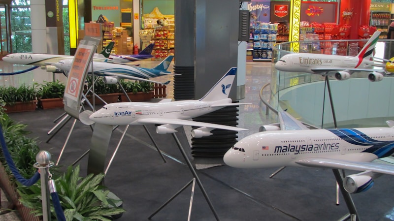 The image features a display of multiple model airplanes situated indoors. Various airline logos are visible on the models, including Malaysia Airlines, Emirates, IranAir, and PIA. The setting appears to be a section of an airport or an aviation-themed exhibit, with a backdrop of colorful retail shops and signage.
