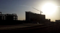 Modern institutional building under construction with cranes and scaffolding at sunrise.