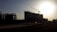 A large construction site with a prominent building structure partially silhouetted against a setting or rising sun. A crane is visible above the building, indicating active construction. The scene is set in an open area with undeveloped land in the foreground and other building structures in the distance.