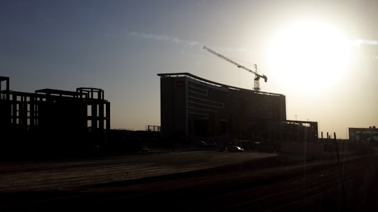 A large construction site with a prominent building structure partially silhouetted against a setting or rising sun. A crane is visible above the building, indicating active construction. The scene is set in an open area with undeveloped land in the foreground and other building structures in the distance.