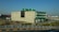 A modern industrial building with a prominent green and beige facade is situated alongside a road. The building features a large green ventilation structure on the roof and is surrounded by a fenced area. The foreground includes some construction debris and landscaped shrubs. In the background, several commercial vehicles and additional industrial structures are visible under a clear blue sky.