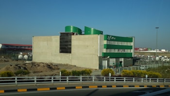 A modern industrial building with a prominent green and beige facade is situated alongside a road. The building features a large green ventilation structure on the roof and is surrounded by a fenced area. The foreground includes some construction debris and landscaped shrubs. In the background, several commercial vehicles and additional industrial structures are visible under a clear blue sky.