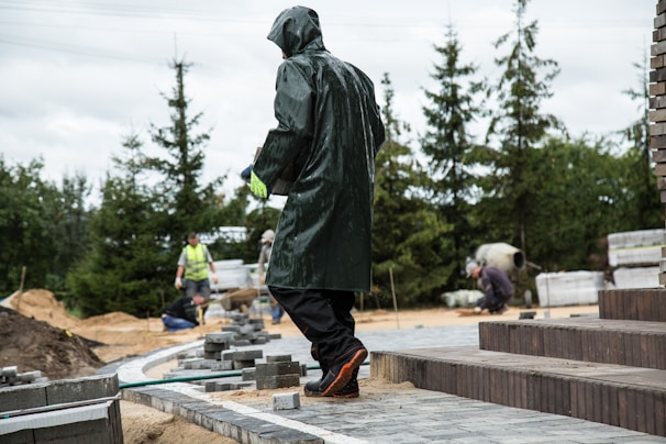 A person in a raincoat is working on a construction site, surrounded by bricks and sand. Other workers are seen in the background, wearing safety gear and performing tasks. The setting includes trees and stacks of materials.