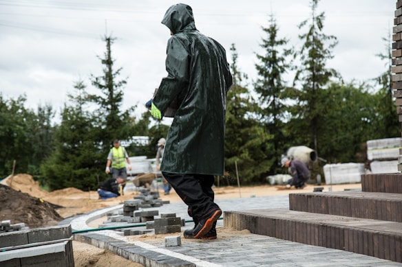 A person in a raincoat is working on a construction site, surrounded by bricks and sand. Other workers are seen in the background, wearing safety gear and performing tasks. The setting includes trees and stacks of materials.
