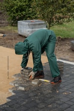 A person wearing green protective clothing and gloves is arranging square paving stones on a partially constructed pathway. The individual is bending over to pick up or place the bricks, which are stacked near their feet. The surroundings include a patch of grass, a wrapped pallet in the background, and a few trees.