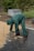 A person wearing green protective clothing and gloves is arranging square paving stones on a partially constructed pathway. The individual is bending over to pick up or place the bricks, which are stacked near their feet. The surroundings include a patch of grass, a wrapped pallet in the background, and a few trees.